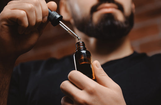 A confident man in his early 30s with a lush beard stands in a stylish bathroom holding a natural herbal hair growth oil.