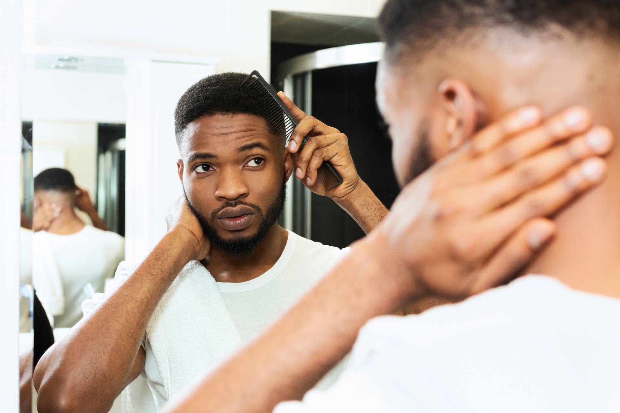 A confident man in his late 20s with well groomed hair and a beard, in a stylish bathroom with grooming products including a comb and leave in conditioner.